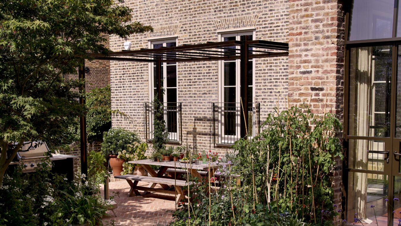 Outdoor patio with a wooden dining table beneath a dark pergola and surrounding greenery