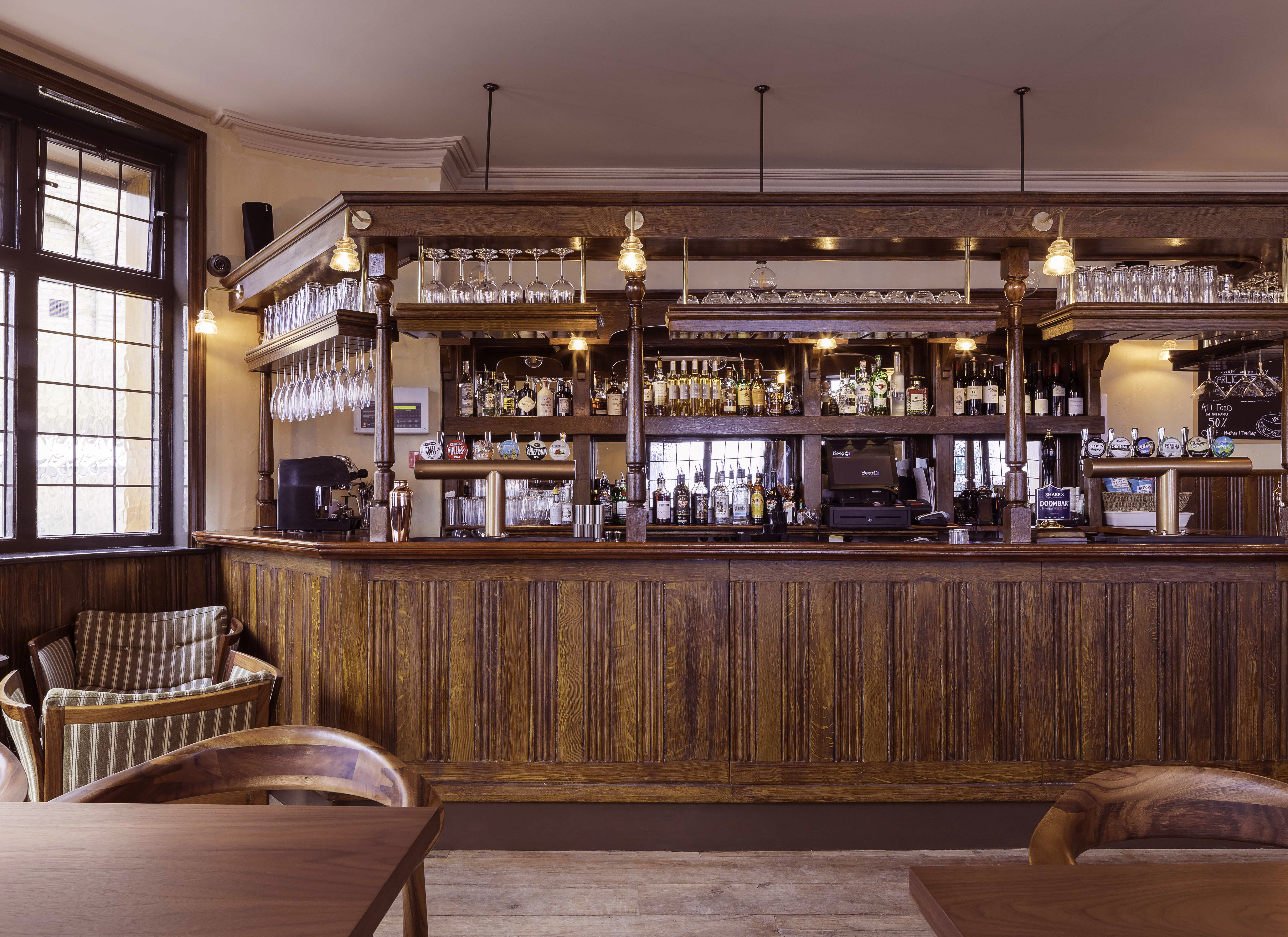 A straight-on interior view of a restored wooden pub bar with intricate paneling, overhead glass racks, and a stocked back bar